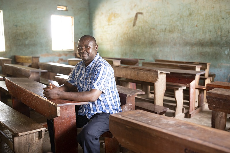 A middle aged man sits in an empty classroom in Uganda looking at the camera.