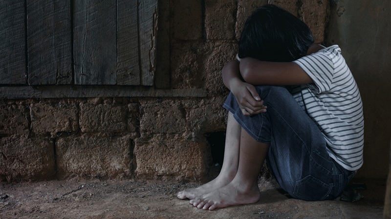 A young girl sits against a wall with her face in her hands.