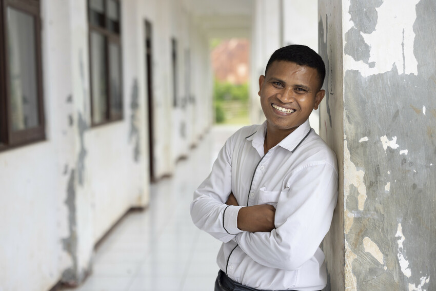 Well-dressed man stands against wall of an Indonesian school, smiling at camera.