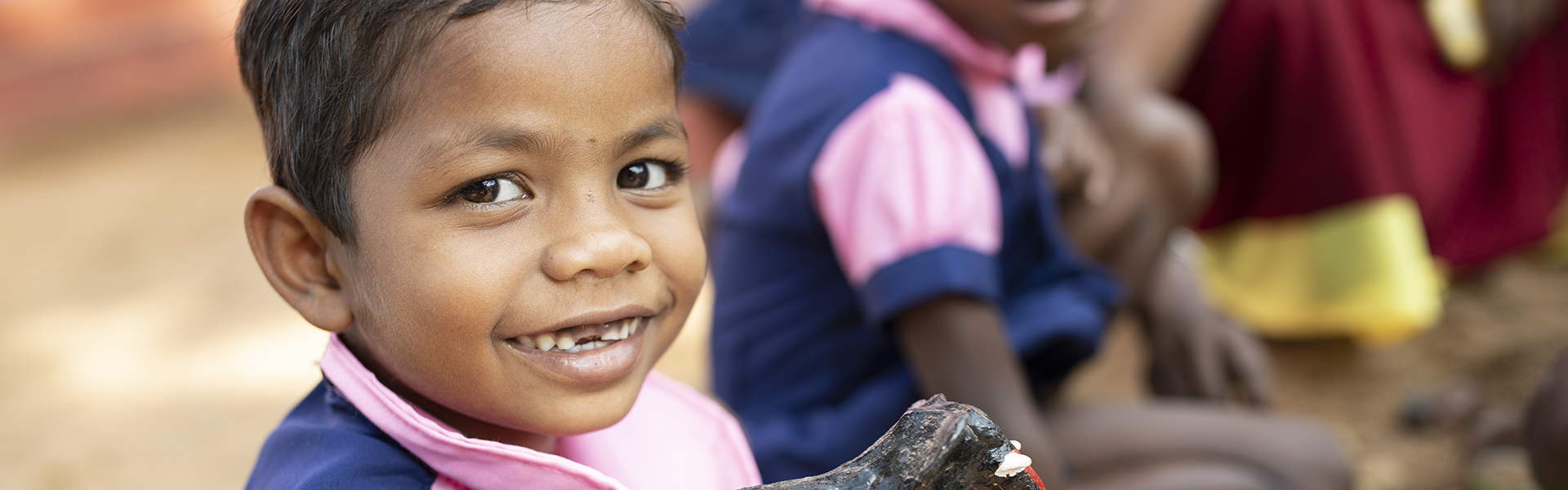 A cheerful young child in a pink and blue school uniform smiles while sitting outside, with other students visible in the background.