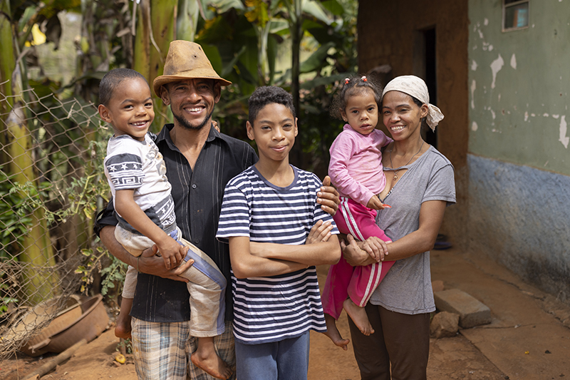 A smiling family of four poses outside their rural home, with the father holding a young boy and the mother holding a little girl.