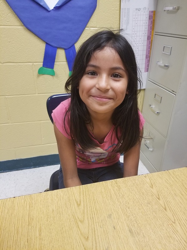 a young girl with dark hair sits at a desk in school