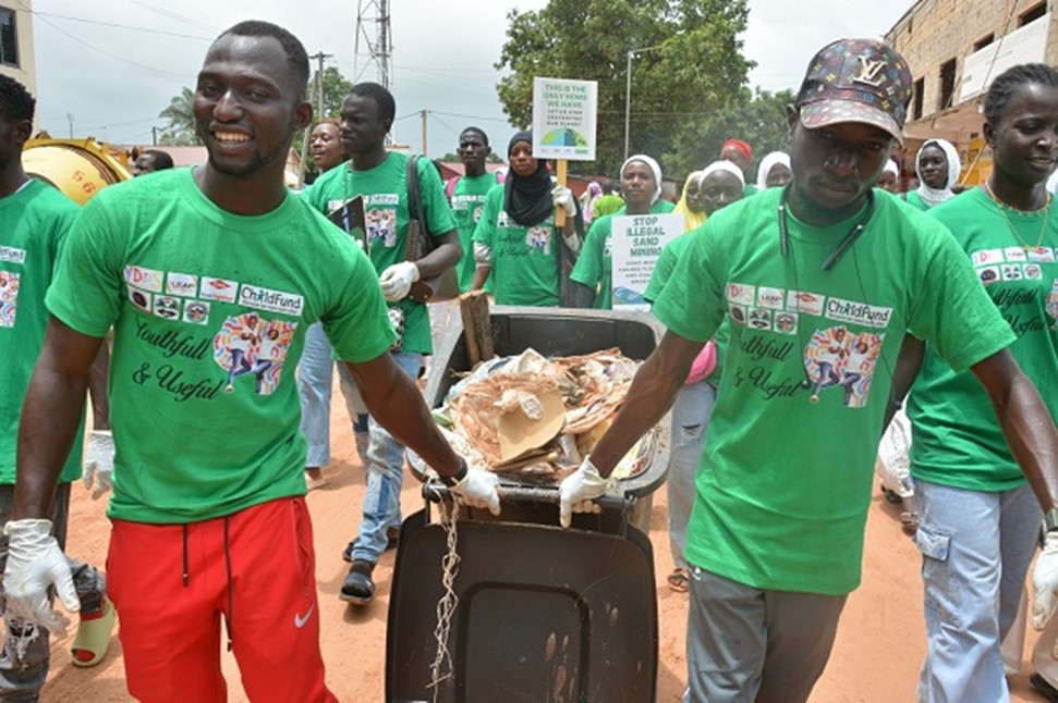 Smiling volunteers in green shirts push a trash cart during a community clean-up event.