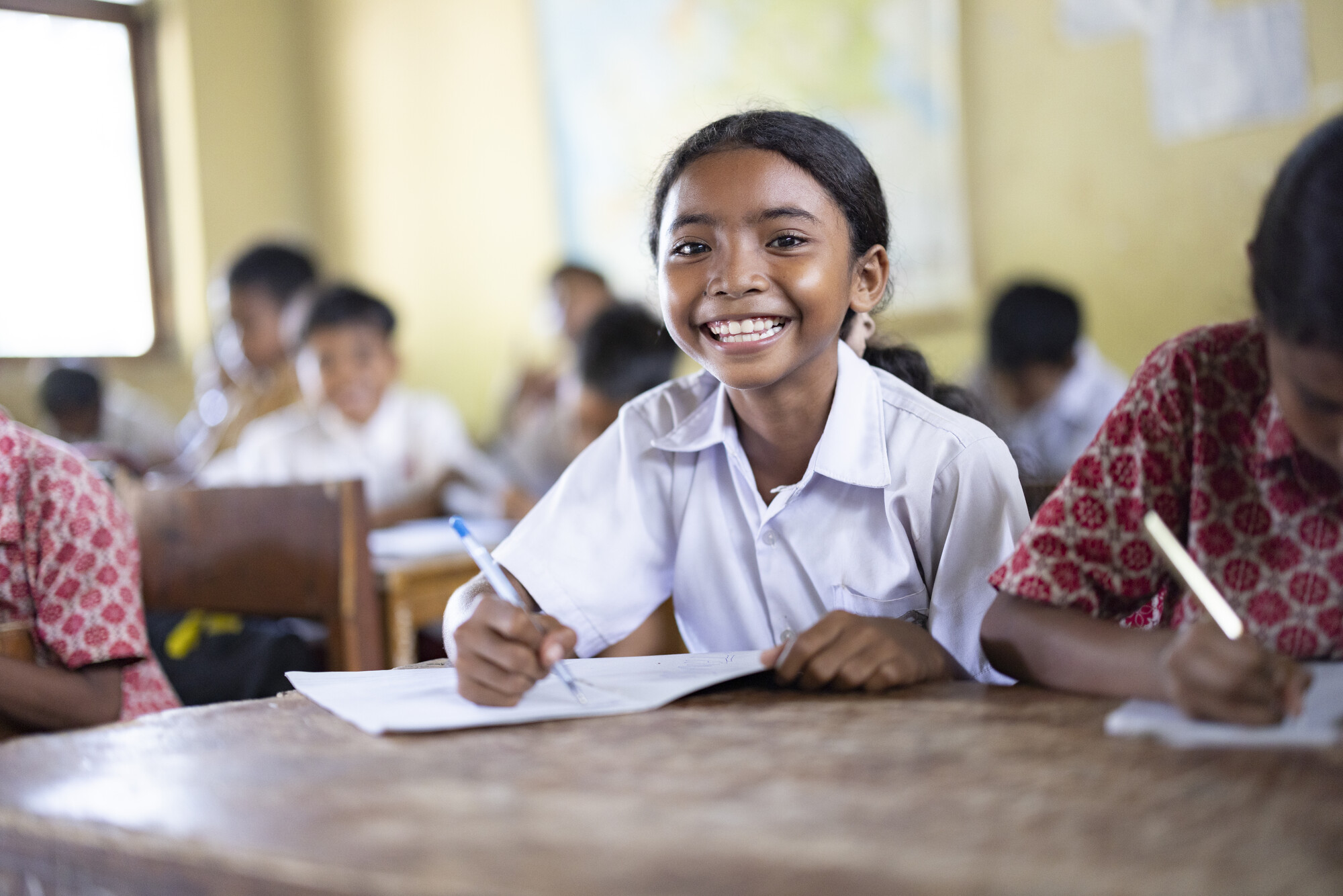 A girl in Indonesia sits at a desk in a classroom, smiling brightly.